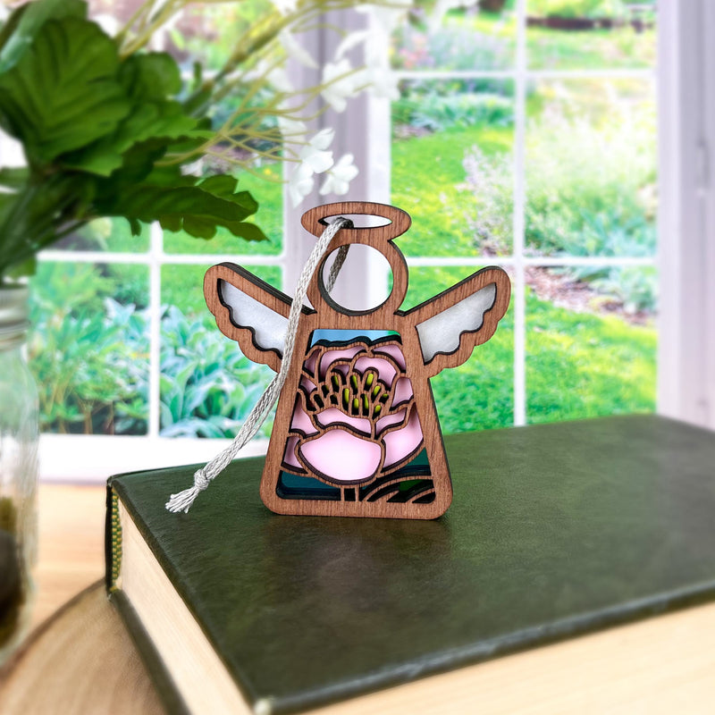 The Forged Flare® Pink Peony Ornament is a 3.5" Mother's Angels® wooden angel figurine with stained glass-style floral details, a white tassel, and green leaves—perfect as a Christmas tree ornament or decorative accent.