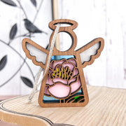 The Forged Flare® Pink Peony Ornament | 3.5" Mother's Angels® Angel Figurine stands on a light wooden surface, with a blurred metal bird wall art and subtly lined white wall in the background. Perfect as a Christmas ornament.