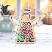 The Forged Flare® Purple Lilac Ornament, a 3.5" Mother's Angels® wooden angel figurine with cut-out wings and a dress featuring purple flowers and green leaves, stands on a white surface with blurred vases and a window in the background.