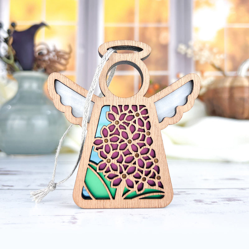 The Forged Flare® Purple Lilac Ornament, a 3.5" Mother's Angels® wooden angel figurine with cut-out wings and a dress featuring purple flowers and green leaves, stands on a white surface with blurred vases and a window in the background.