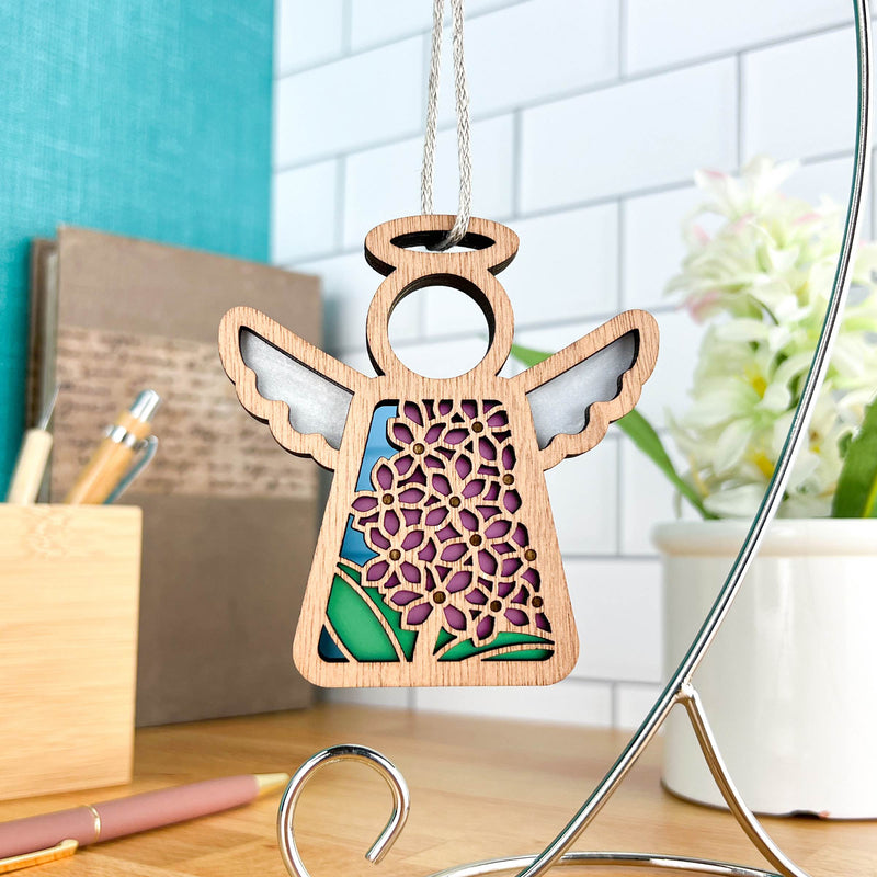 The Purple Lilac Ornament, a 3.5" angel figurine by Forged Flare® from Mother's Angels®, features intricate floral cutouts and stained-glass effects. Displayed on a stand, it makes a charming decor piece near your desk or as a Christmas tree ornament.