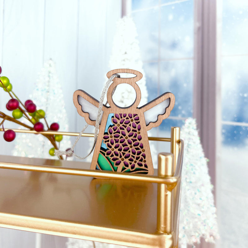 The Forged Flare® Purple Lilac Ornament | 3.5" (H) Mother's Angels® Angel Figurine, with floral cutouts, hangs on a gold shelf amid snowy trees, red berries, and a frosted window for a festive winter holiday display.