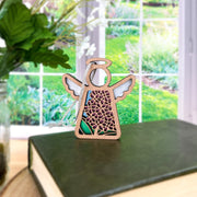 The Forged Flare® Mother's Angels® Purple Lilac Ornament is a 3.5" wooden angel figurine with floral cut-outs, displayed on a green book near a window with garden views. A glass jar with leaves adds to its Christmas ornament charm.
