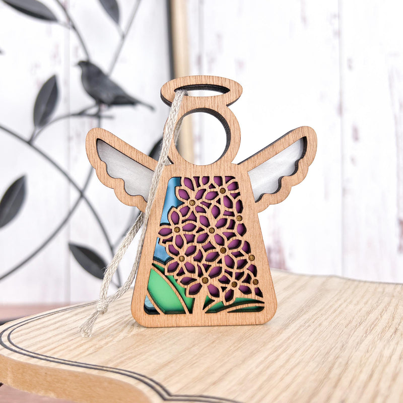 The Forged Flare® Purple Lilac Ornament is a 3.5" Mother's Angels® wooden angel figurine with floral cutouts and purple-green accents, complete with a hanging string—ideal as a Christmas tree ornament. A metal bird and white wall appear blurred in the background.