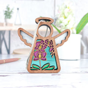 The Forged Flare® Purple Orchid Ornament, a 3.5" Mother's Angels® angel figurine, with silver wings, a halo, and colorful floral design on turquoise, sits on a white surface; blurred plants and desk items appear in the background.
