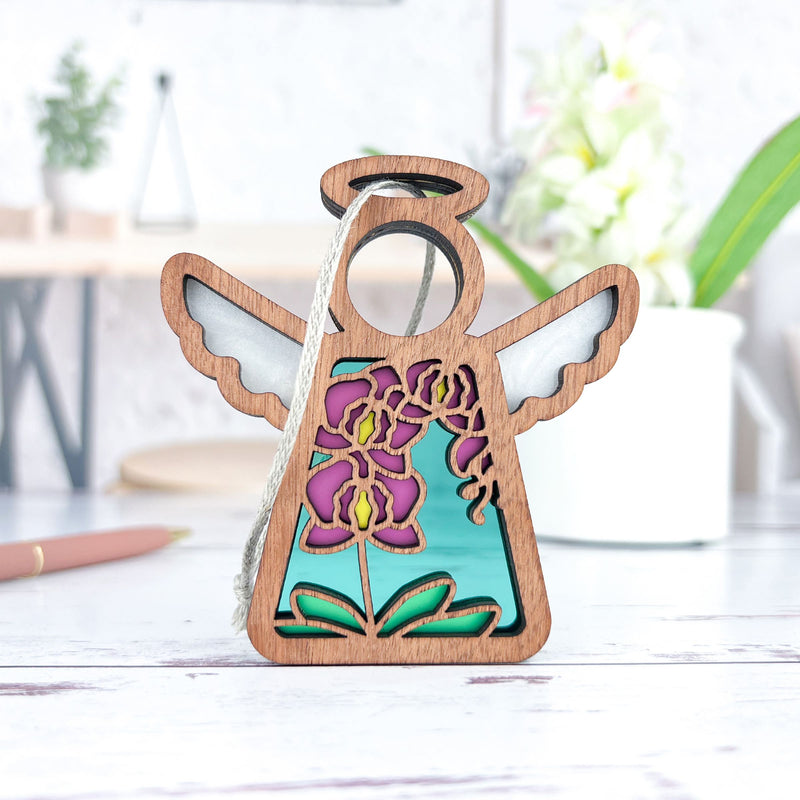 The Forged Flare® Purple Orchid Ornament, a 3.5" Mother's Angels® angel figurine, with silver wings, a halo, and colorful floral design on turquoise, sits on a white surface; blurred plants and desk items appear in the background.