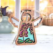 The Forged Flare® Purple Orchid Ornament, a 3.5" tall Mother's Angels® angel figurine with cutout wings and a turquoise background, displays colorful floral designs as it sits on a white surface with blurred vases and autumn scenery behind.