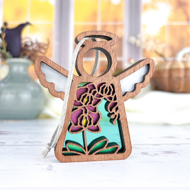 The Forged Flare® Purple Orchid Ornament, a 3.5" tall Mother's Angels® angel figurine with cutout wings and a turquoise background, displays colorful floral designs as it sits on a white surface with blurred vases and autumn scenery behind.