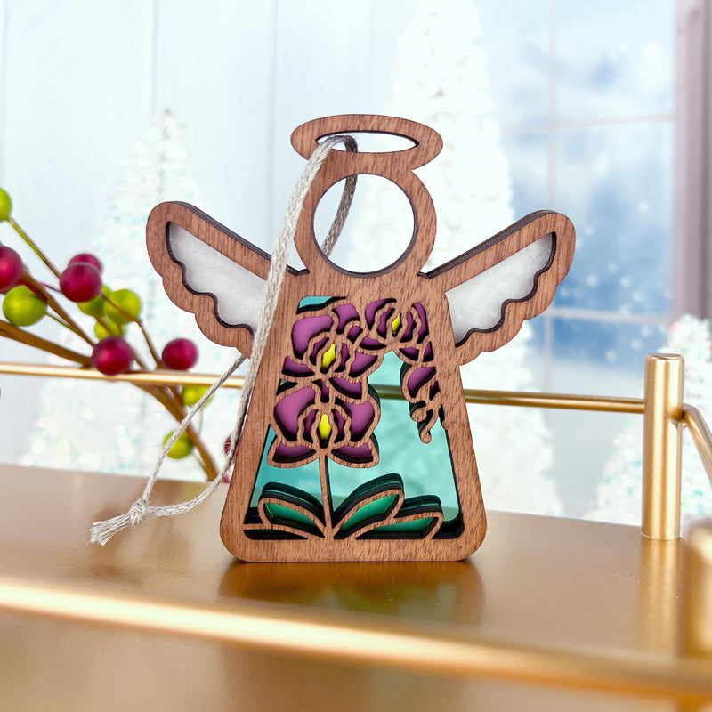The Forged Flare® Purple Orchid Ornament is a 3.5" Mother's Angels® angel figurine, featuring purple and yellow floral designs, white ribbon, and displayed on a gold stand with frosted trees and a snowy window backdrop.