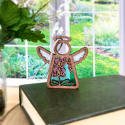 The Forged Flare® Purple Orchid Ornament, a 3.5" Mother's Angels® angel figurine, rests on a green book as sunlight fills the room, with a glass vase of white flowers nearby and garden views through white-paned windows.