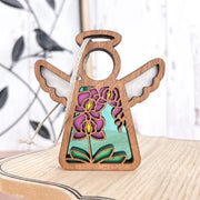 The Forged Flare® Mother's Angels® Purple Orchid Ornament is a 3.5" wooden angel with cut-out wings and a stained glass-style body, ideal for collections or holiday décor, shown on light wood with leafy black metal accents behind.