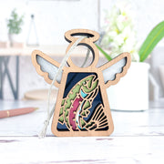The Forged Flare® Mother's Angels® 3.5" angel figurine features a rainbow trout fish ornament cutout, silver wings, and a hanging loop, displayed on a light tabletop with a blurred background.