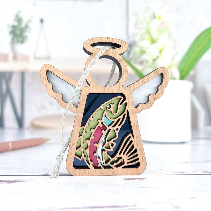 The Forged Flare® Mother's Angels® 3.5" angel figurine features a rainbow trout fish ornament cutout, silver wings, and a hanging loop, displayed on a light tabletop with a blurred background.