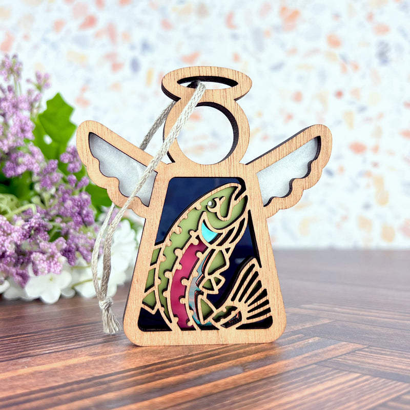 The Forged Flare® Mother's Angels® 3.5" Rainbow Trout Fish Ornament features a wooden angel figurine with a trout cutout and a silver string, ideal for use as a Christmas tree ornament or decor. Purple flowers and a speckled backdrop add charm.