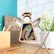 The Forged Flare® Mother's Angels® 3.5" wooden angel figurine features a rainbow trout fish cutout and hangs on a string, shown against a white tile wall with books, pens, and a potted plant on a wooden desk.