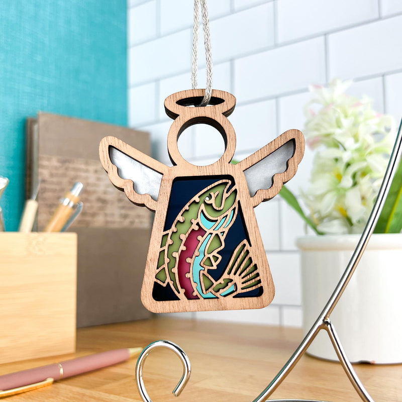 The Forged Flare® Mother's Angels® 3.5" wooden angel figurine features a rainbow trout fish cutout and hangs on a string, shown against a white tile wall with books, pens, and a potted plant on a wooden desk.
