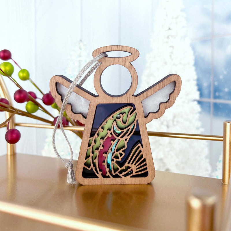 The Forged Flare® Rainbow Trout Fish Ornament is a 3.5" Mother's Angels® wooden figurine featuring silver wings and a vibrant trout design, perfect for festive shelf displays with holiday decor and snowy trees.