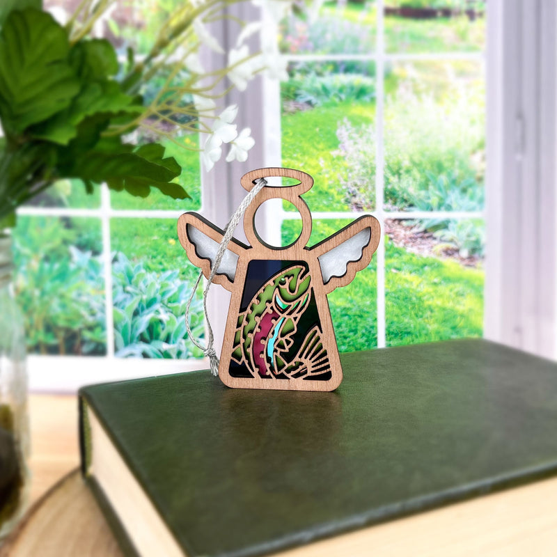 A Forged Flare® Mother's Angels® 3.5" angel figurine with a Rainbow Trout Fish Ornament sits on a green book, with sunlight streaming from a garden window in the background.