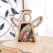 The Forged Flare® Mother's Angels® 3.5" wooden angel figurine features a vibrant rainbow trout fish ornament cutout in the center and stands on a wooden surface, with a metal bird decoration and white wall behind it.