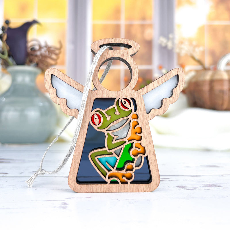 The Forged Flare® Red-Eyed Tree Frog Ornament is a 3.5" Mother's Angels® angel figurine with white wings and a vibrant frog design, perfect as a charming frog gift. It's displayed on a light table with vases and a window blurred in the background.