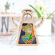 The Red-Eyed Tree Frog Ornament by Forged Flare® is a 3.5" H Mother's Angels® figurine featuring a wooden angel with white wings and a colorful frog, perfect for your Christmas tree or as a unique gift. Hangs by string; background includes blurred plants and objects.