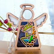 The Forged Flare® Red-Eyed Tree Frog Ornament is a 3.5" wooden angel figurine featuring a vibrant frog design and white wings, perfect for Christmas trees or as a unique gift. It hangs by a white string with festive berry accents in the background.