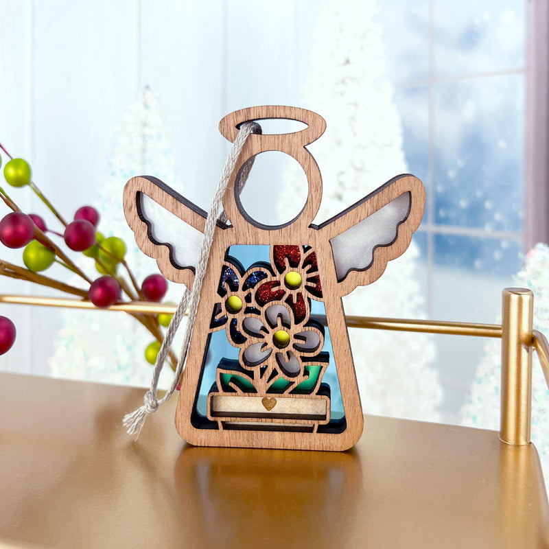 The Forged Flare® Red, White & Bloom Ornament is a 3.5" Mother's Angels® figurine featuring floral designs and gold accents, hanging by a silver string on a gold rail amid red berries and snowy trees—perfect for your Christmas tree.