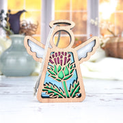 The Forged Flare® Scotch Thistle Ornament is a 3.5" Mother's Angels® figurine featuring a wooden angel with wings, halo, and a green thistle cut-out set against a blue sky, displayed on a white surface with blurred vases in the background.