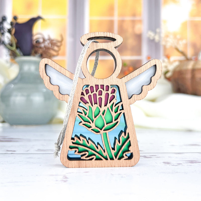 The Forged Flare® Scotch Thistle Ornament is a 3.5" Mother's Angels® figurine featuring a wooden angel with wings, halo, and a green thistle cut-out set against a blue sky, displayed on a white surface with blurred vases in the background.
