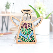 The Forged Flare® Scotch Thistle Ornament is a 3.5" Mother's Angels® wooden angel figurine with cutout wings, a halo, and a colorful thistle flower design symbolizing resilience, displayed on a white surface with green plants in the background.