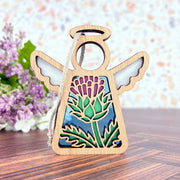 The Forged Flare® Scotch Thistle Ornament is a 3.5" Mother's Angels® figurine featuring stained glass-style wings and a thistle design, shown on wood with purple flowers in the background, reflecting thistle symbolism.