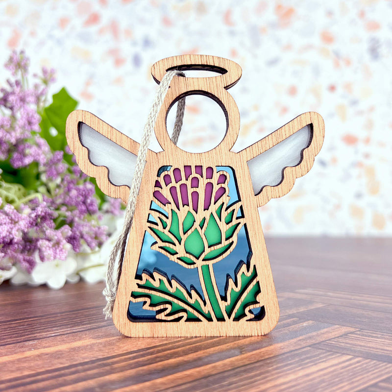 The Forged Flare® Scotch Thistle Ornament is a 3.5" Mother's Angels® figurine featuring stained glass-style wings and a thistle design, shown on wood with purple flowers in the background, reflecting thistle symbolism.