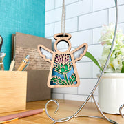 The Scotch Thistle Ornament by Forged Flare® is a 3.5" wooden angel figurine with stained glass-style thistle wings and halo, from the Mother's Angels® line. It hangs on a silver stand amid pens, notebooks, and white flowers.
