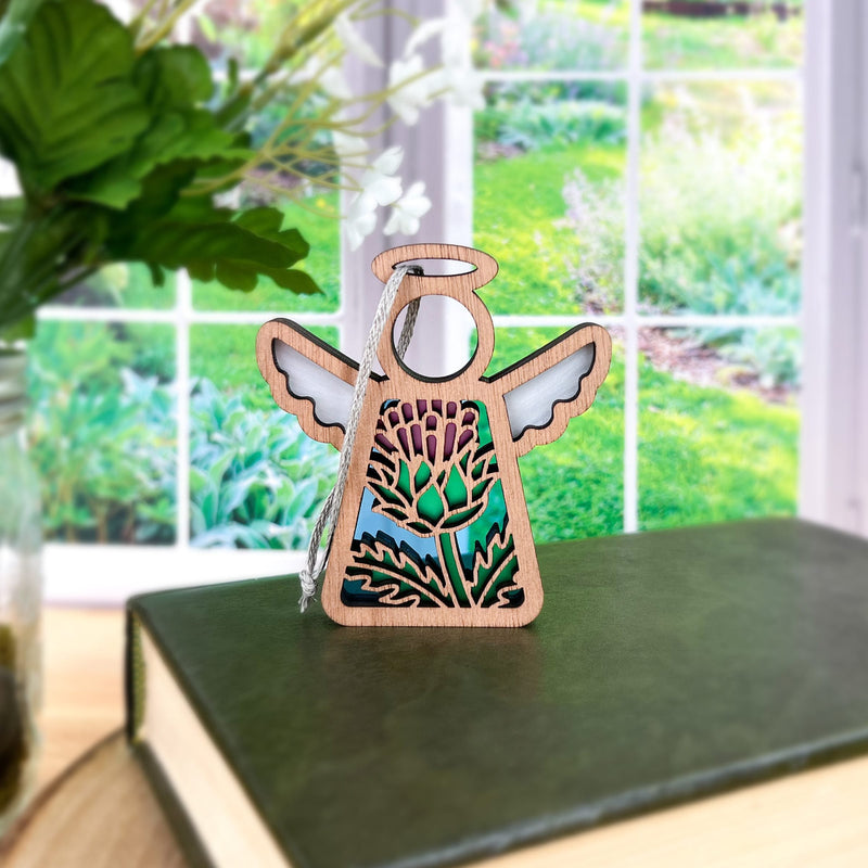 The Forged Flare® Scotch Thistle Ornament, a 3.5" Mother's Angels® figurine, features stained glass wings and floral details as it stands on a green book by the window, looking out at a vibrant garden.