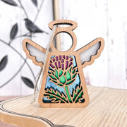 The Forged Flare® Scotch Thistle Ornament, a 3.5" Mother’s Angels® wooden angel figurine with cut-out wings and a colorful thistle design, stands on light wood with leafy metal décor in the background.