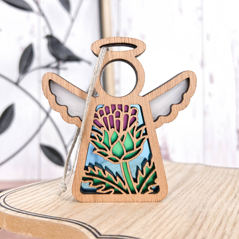 The Forged Flare® Scotch Thistle Ornament, a 3.5" Mother’s Angels® wooden angel figurine with cut-out wings and a colorful thistle design, stands on light wood with leafy metal décor in the background.