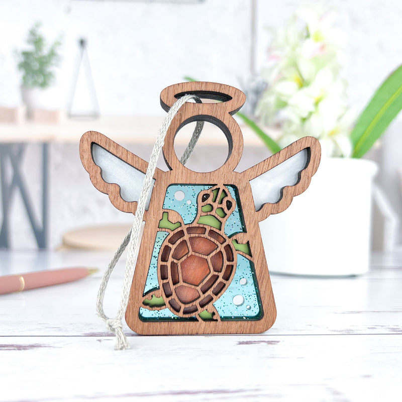 The Sea Turtle Ornament by Forged Flare® is a 3.5" Mother's Angels® figurine featuring a wooden angel with white wings, a halo, and a turtle motif. It hangs by a string against a blue background—ideal beach house decor.