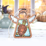 The Forged Flare® Sea Turtle Ornament, a 3.5" Mother's Angels® angel figurine with a blue and green stained-glass style sea turtle design, stands on a white surface—ideal beach house decor. Blurred vases and autumn accents appear in the background.