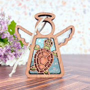 The Forged Flare® Sea Turtle with Imperfections Ornament, a 3.5" Mother's Angels® figurine, features a turtle cutout design—ideal as coastal decor or a Christmas ornament. Displayed on wood with purple flowers in the background.