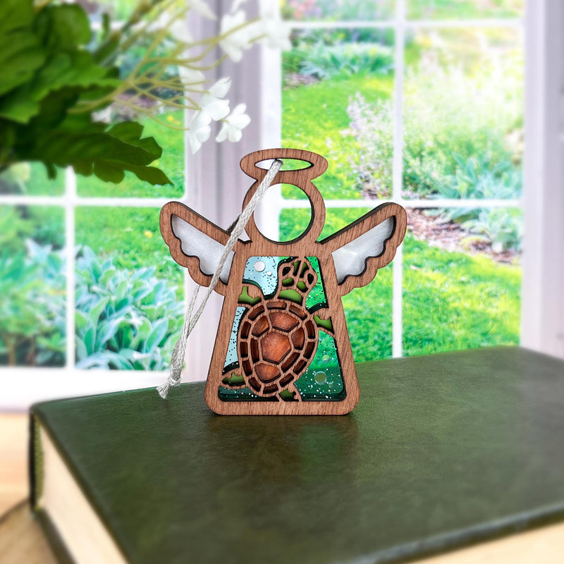 The Forged Flare® Mother's Angels® Sea Turtle with Imperfections Ornament, a 3.5" wooden angel figurine, rests on a closed green book by a sunlit window with lush greenery—perfect as a unique Christmas tree ornament.