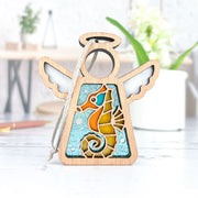 The Seahorse Ornament by Forged Flare® is a 3.5" wooden angel figurine from Mother's Angels®, featuring stained glass-style seahorse art—ideal as a Christmas tree ornament or unique beach house decor.