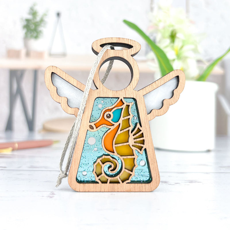 The Seahorse Ornament by Forged Flare® is a 3.5" wooden angel figurine from Mother's Angels®, featuring stained glass-style seahorse art—ideal as a Christmas tree ornament or unique beach house decor.