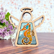 The Forged Flare® Seahorse Ornament—Mother's Angels® 3.5" Angel Figurine—features stained glass-style wings and a seahorse design, ideal as a Christmas tree ornament or beach house decor. Seen on wood with purple flowers and a colorful background.