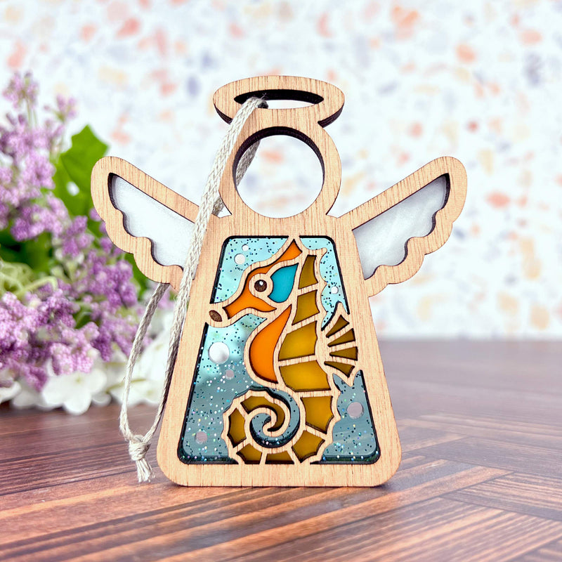 The Forged Flare® Seahorse Ornament—Mother's Angels® 3.5" Angel Figurine—features stained glass-style wings and a seahorse design, ideal as a Christmas tree ornament or beach house decor. Seen on wood with purple flowers and a colorful background.