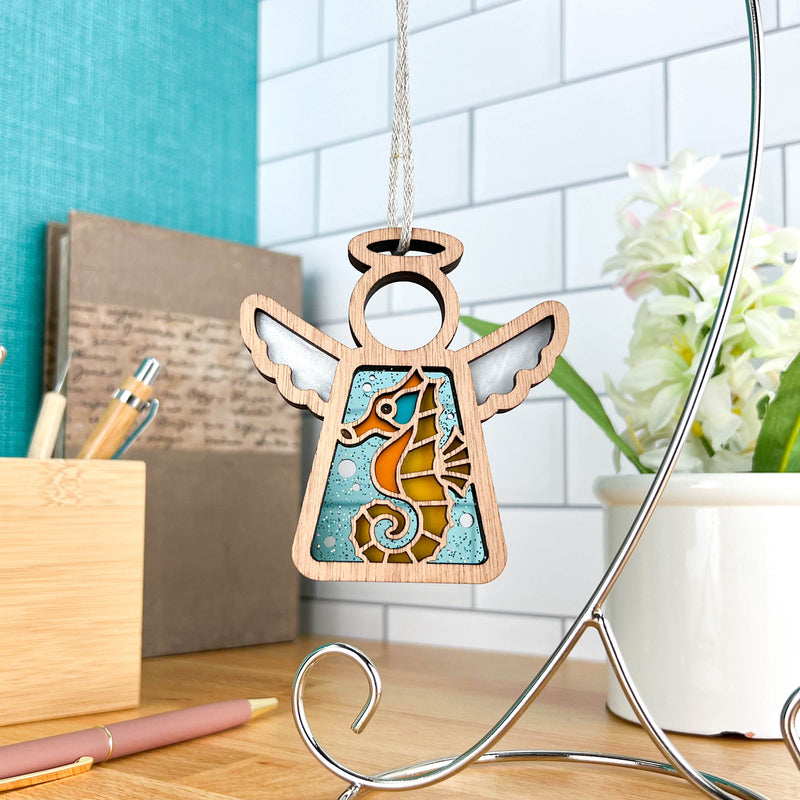 The Forged Flare® Seahorse Ornament (3.5" Mother's Angels® Angel Figurine) hangs from a silver stand on a desk—ideal for beach house decor or as a distinctive Christmas ornament, with office supplies and a potted plant in the background.