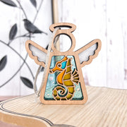 The Forged Flare® Seahorse Ornament | 3.5" (H) Angel Figurine from Mother's Angels® features a stained glass blue and orange seahorse, ideal as a unique Christmas tree ornament or beach house decor; blurred bird-shaped wall art in background.