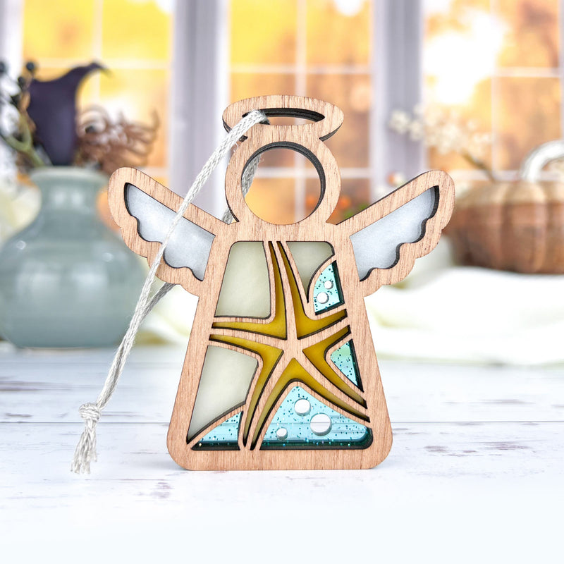 The Forged Flare® Starfish Ornament is a 3.5" Mother's Angels® figurine with wings, halo, stained glass, and blue accents—a perfect beach house decor piece—displayed on a white surface amid autumn décor and windows.