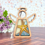 The Starfish Ornament, a 3.5" wooden angel figurine by Forged Flare® Mother's Angels®, features yellow, cream, and teal stained glass—ideal for beach house decor. It stands on wood with blurred purple flowers and greenery in the background.