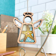 The Forged Flare® Mother's Angels® Starfish Ornament is a 3.5" angel figurine with a stained glass effect and yellow starfish design—perfect for beach house decor. A desk, pen, plant, and white tiled wall appear in the background.