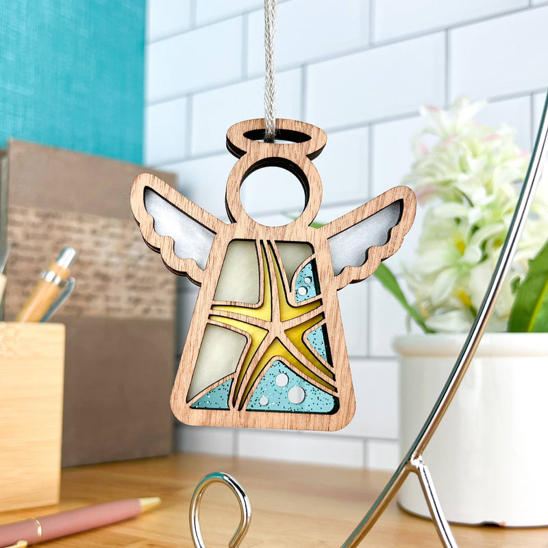 The Forged Flare® Mother's Angels® Starfish Ornament is a 3.5" angel figurine with a stained glass effect and yellow starfish design—perfect for beach house decor. A desk, pen, plant, and white tiled wall appear in the background.